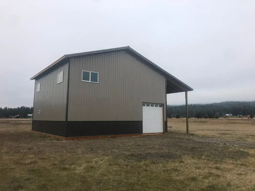 Two-story tan metal building with a white garage door and a small porch. Set in a field on a cloudy day.