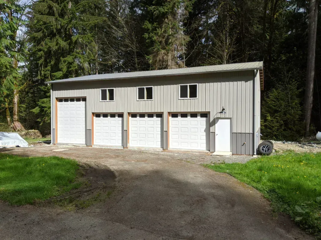 A gray metal garage with three garage doors and a side door, set in a grassy area with trees in the background.