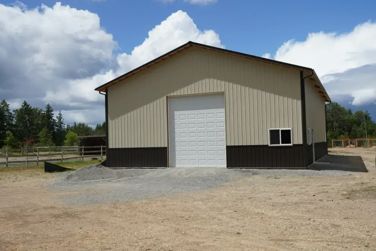 Tan and brown metal building with a white garage door and a small window, on a gravel lot.