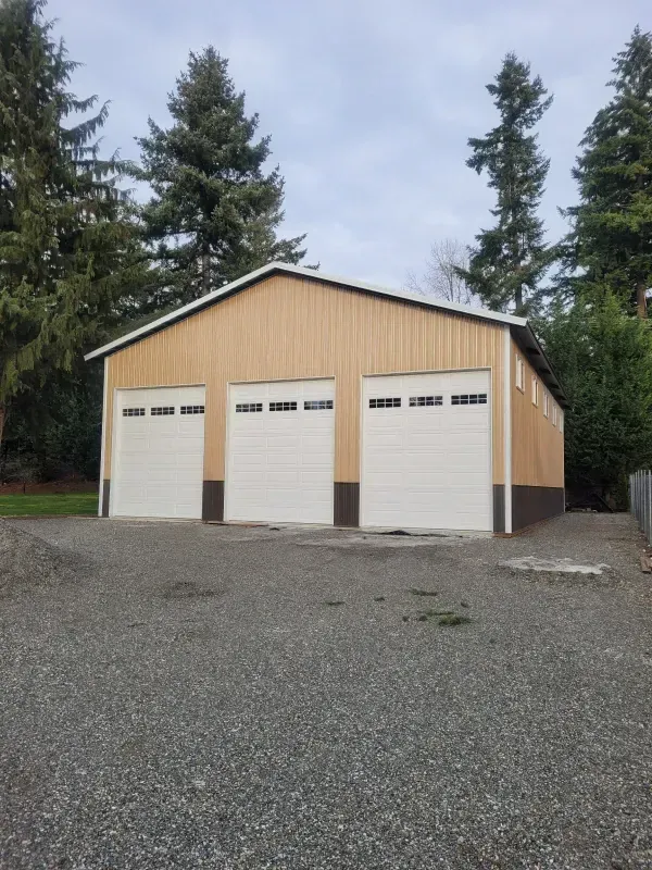 Tan and white three-bay garage with brown trim, gravel driveway, and trees against a cloudy sky.