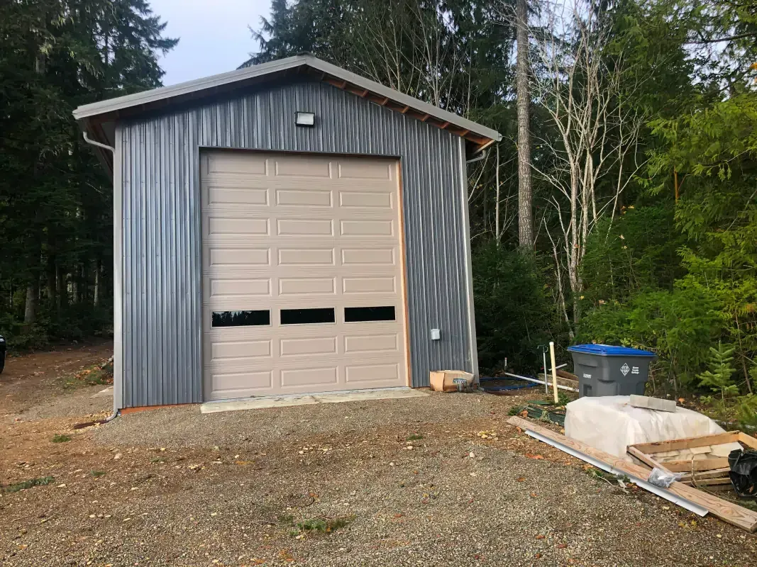 Metal garage with a tan garage door on gravel, set in a forest.