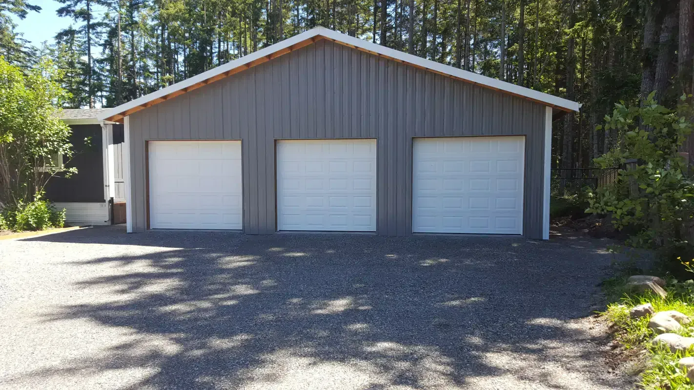 Three-car garage with white doors, gray siding, gravel driveway, and trees in the background.