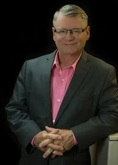 A man in a suit and pink shirt is standing with his hands folded.