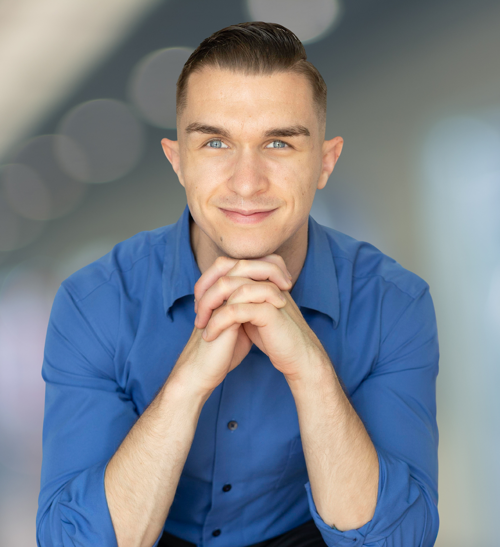 Man in blue shirt, hands clasped under chin, smiling, blurred background.