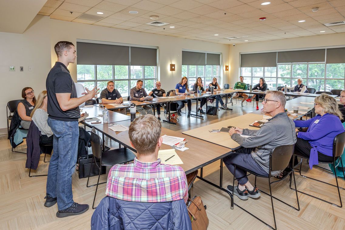 A man presents to a group seated at a long table in a conference room.