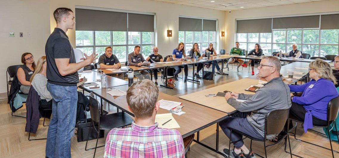 Man with short hair, clasped hands under chin, smiles at camera. Dark polo shirt, natural light.