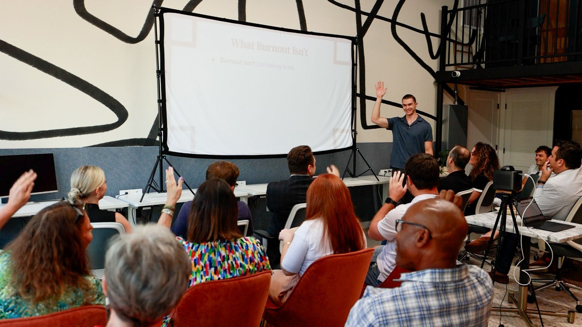 A man reading from papers, a woman using a laptop. Both are seated at tables in a room with neutral walls.