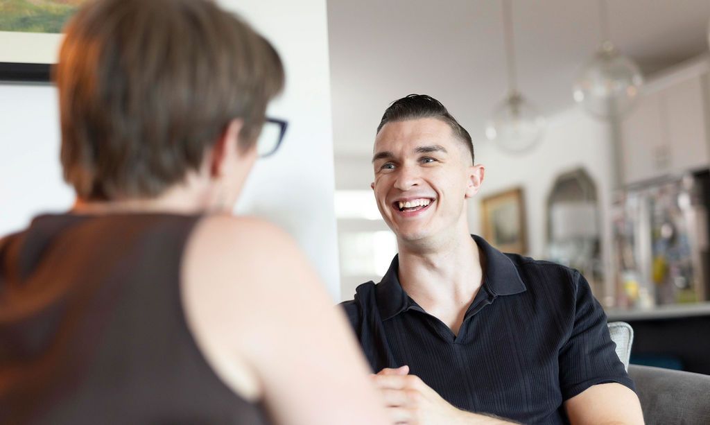 A man with short, dark hair smiles at a person off-camera. He wears a black shirt in a brightly lit room.