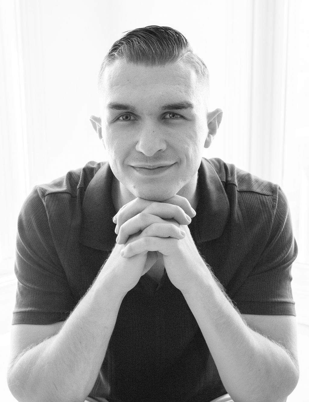 A man with short hair in a dark polo shirt rests his chin on clasped hands, smiling at the camera.