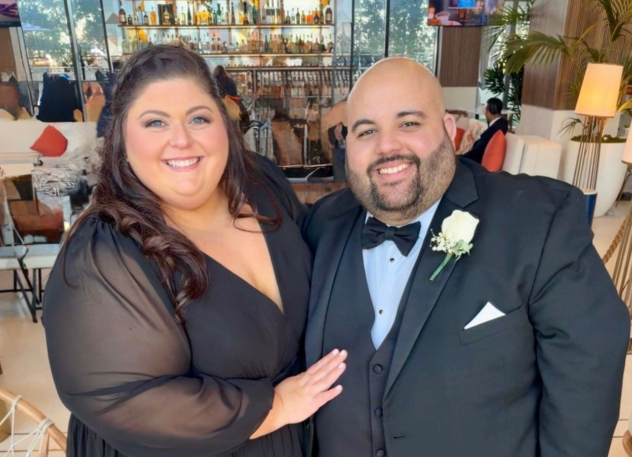 Woman in black dress and man in a suit pose together, smiling, near a bar.