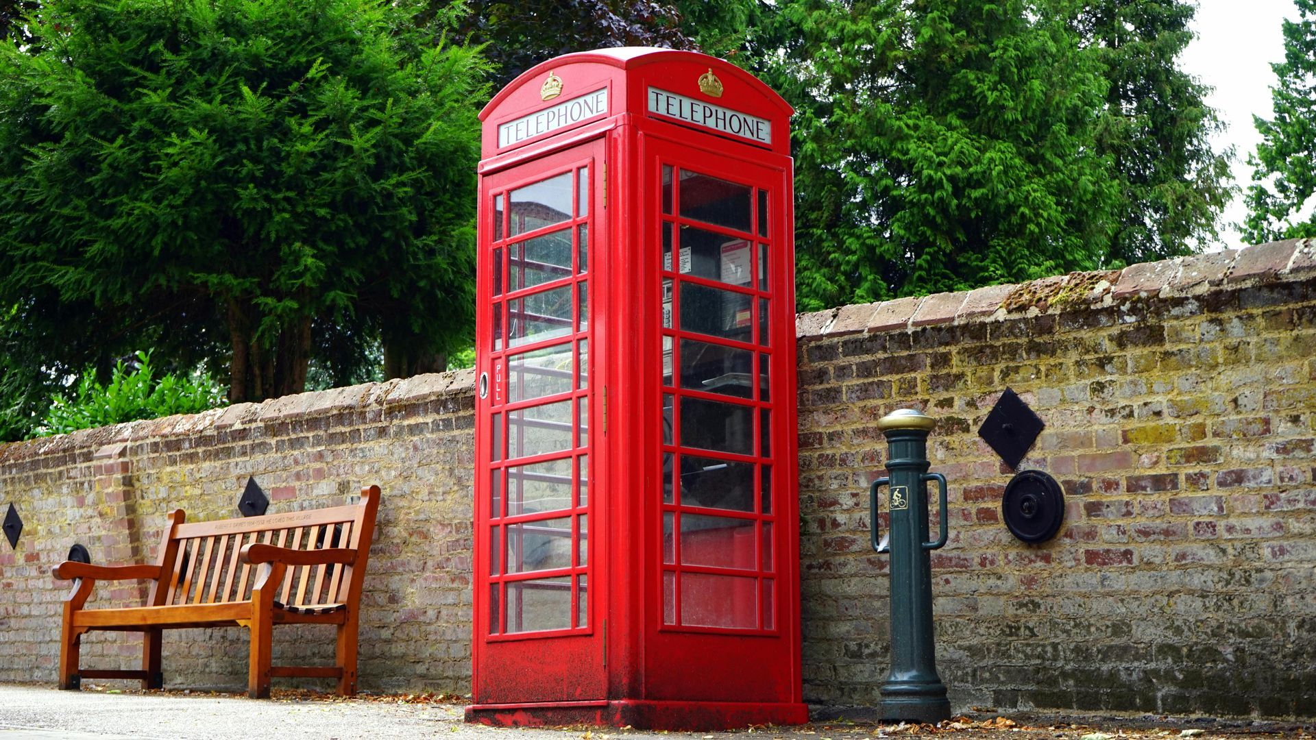 Red British telephone booth next to a brick wall with a wooden bench and greenery in the background.
