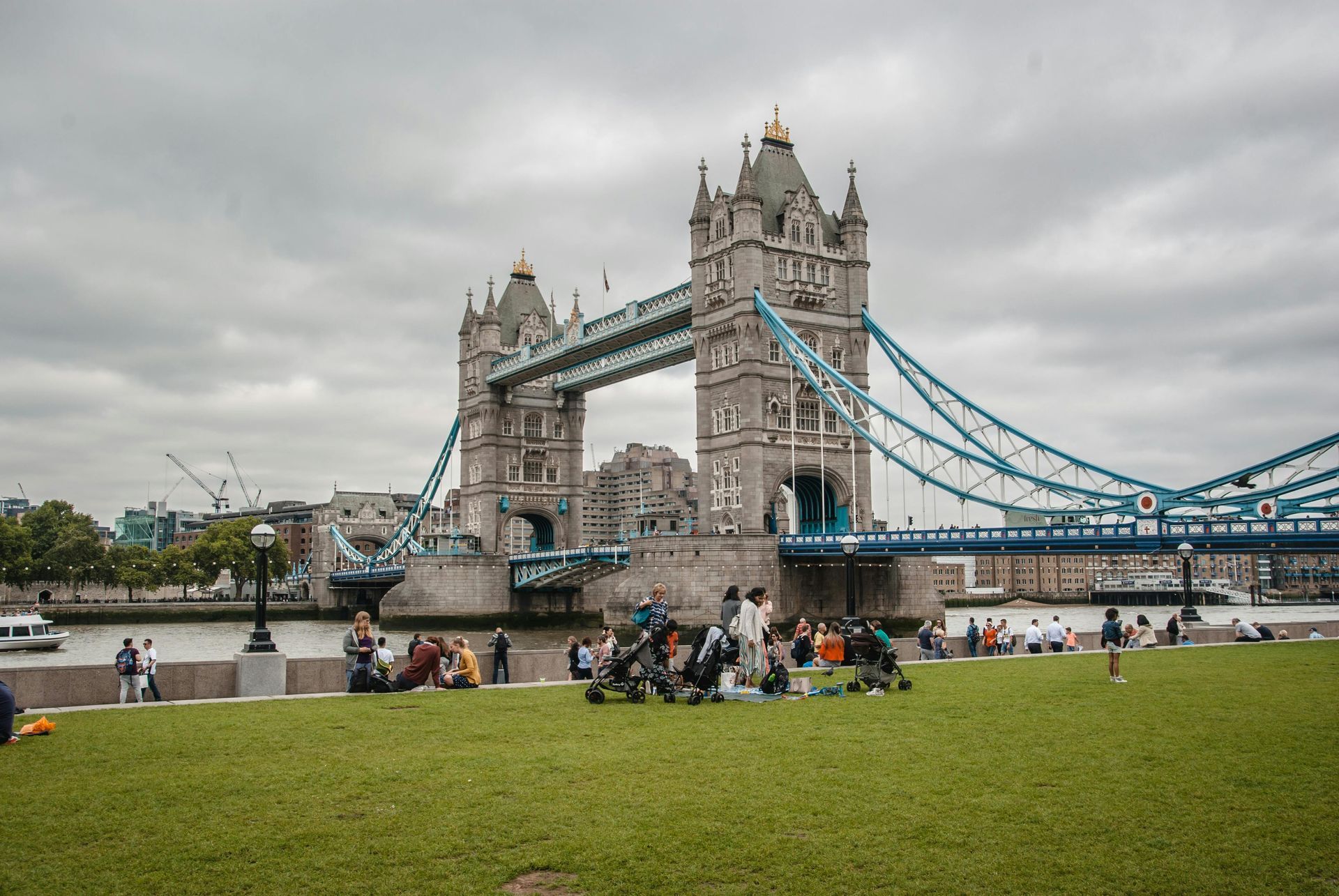 Tower Bridge in London on a cloudy day, with people on a grassy area in the foreground.