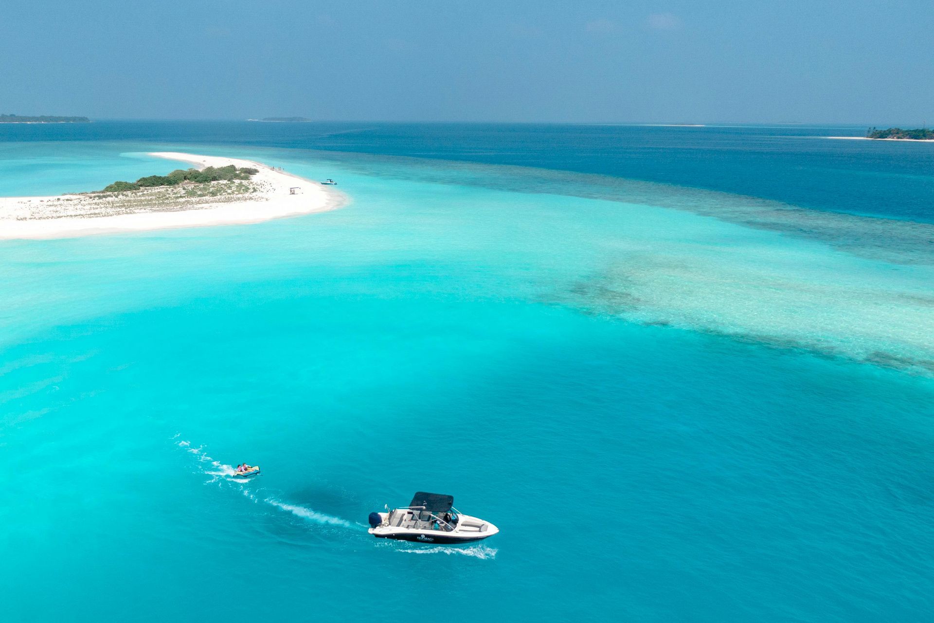 Boat sails through turquoise water toward a small, sandy island under a clear blue sky.