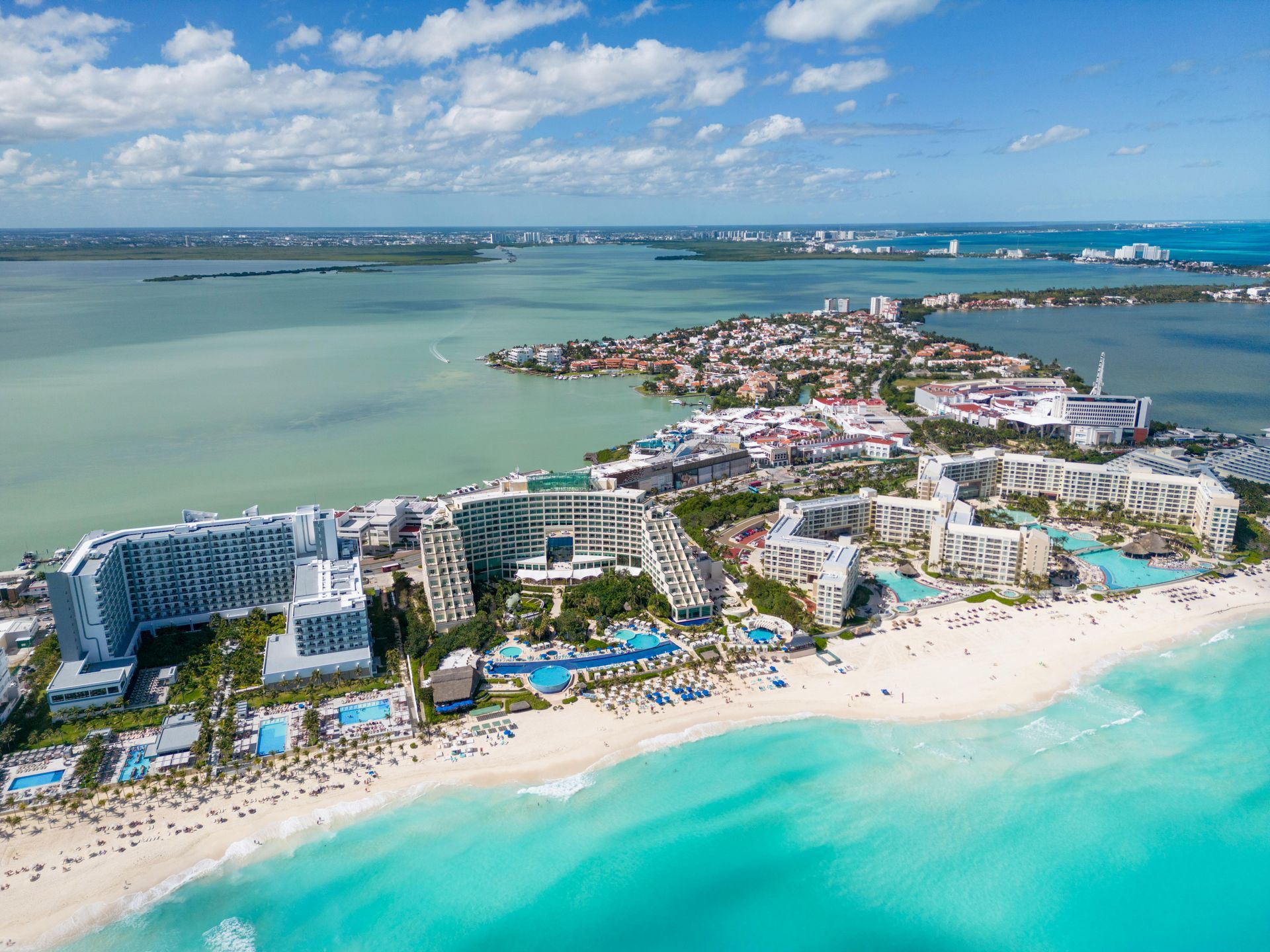 Aerial view of Cancún's hotel zone on a sandy beach with turquoise water. Buildings, pools, and a lagoon in the background.