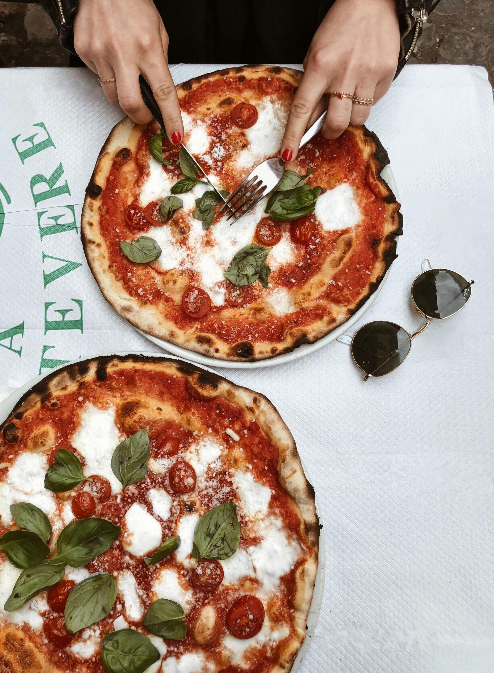Two Margherita pizzas on a white tablecloth; person's hands using a fork and knife.
