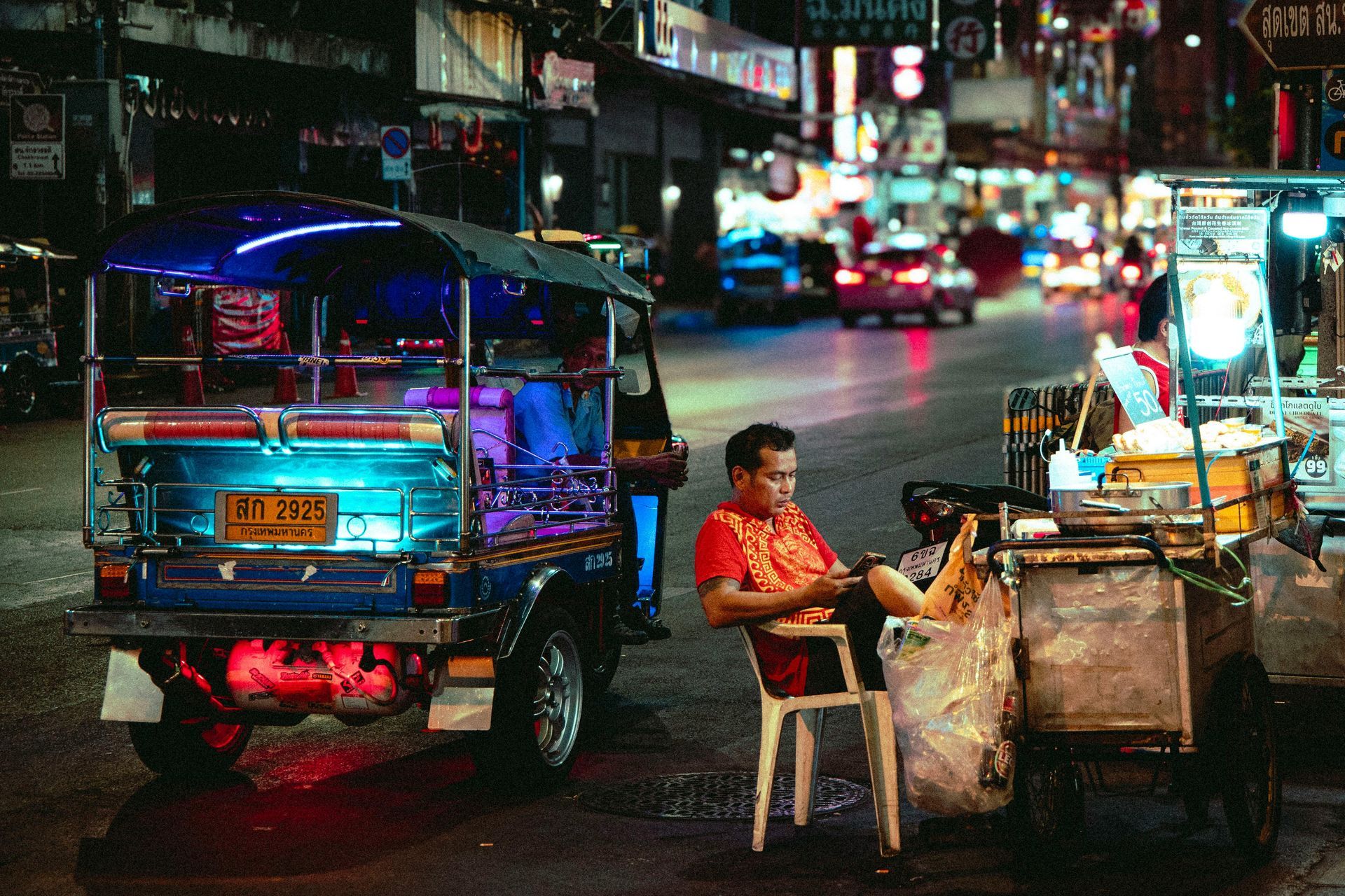 Tuk-tuk driver sits on a chair next to his vehicle, illuminated by neon lights on a city street at night.