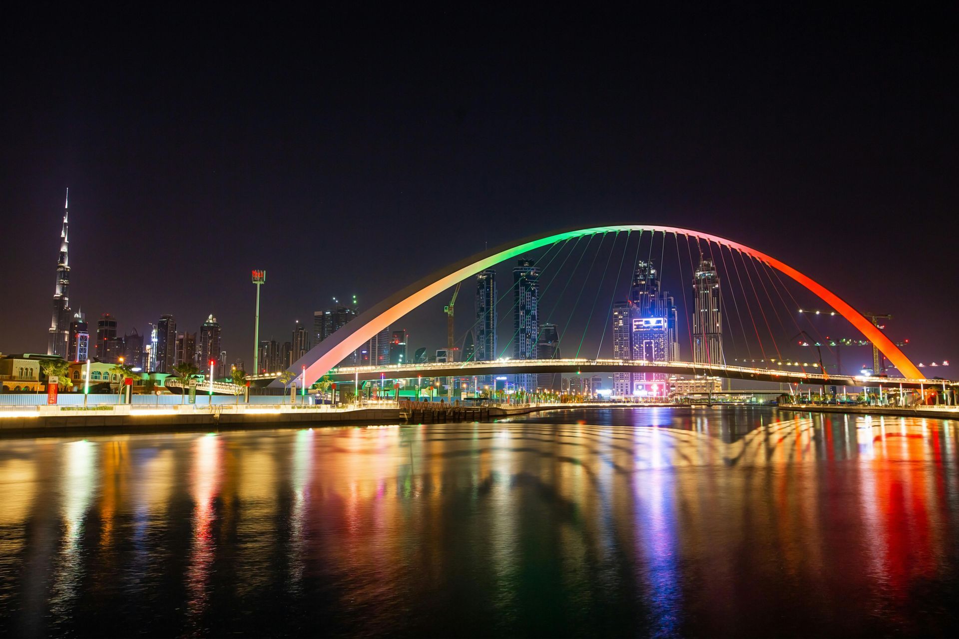 Night view of Dubai Water Canal, illuminated arch bridge with rainbow lighting, skyline in the background.