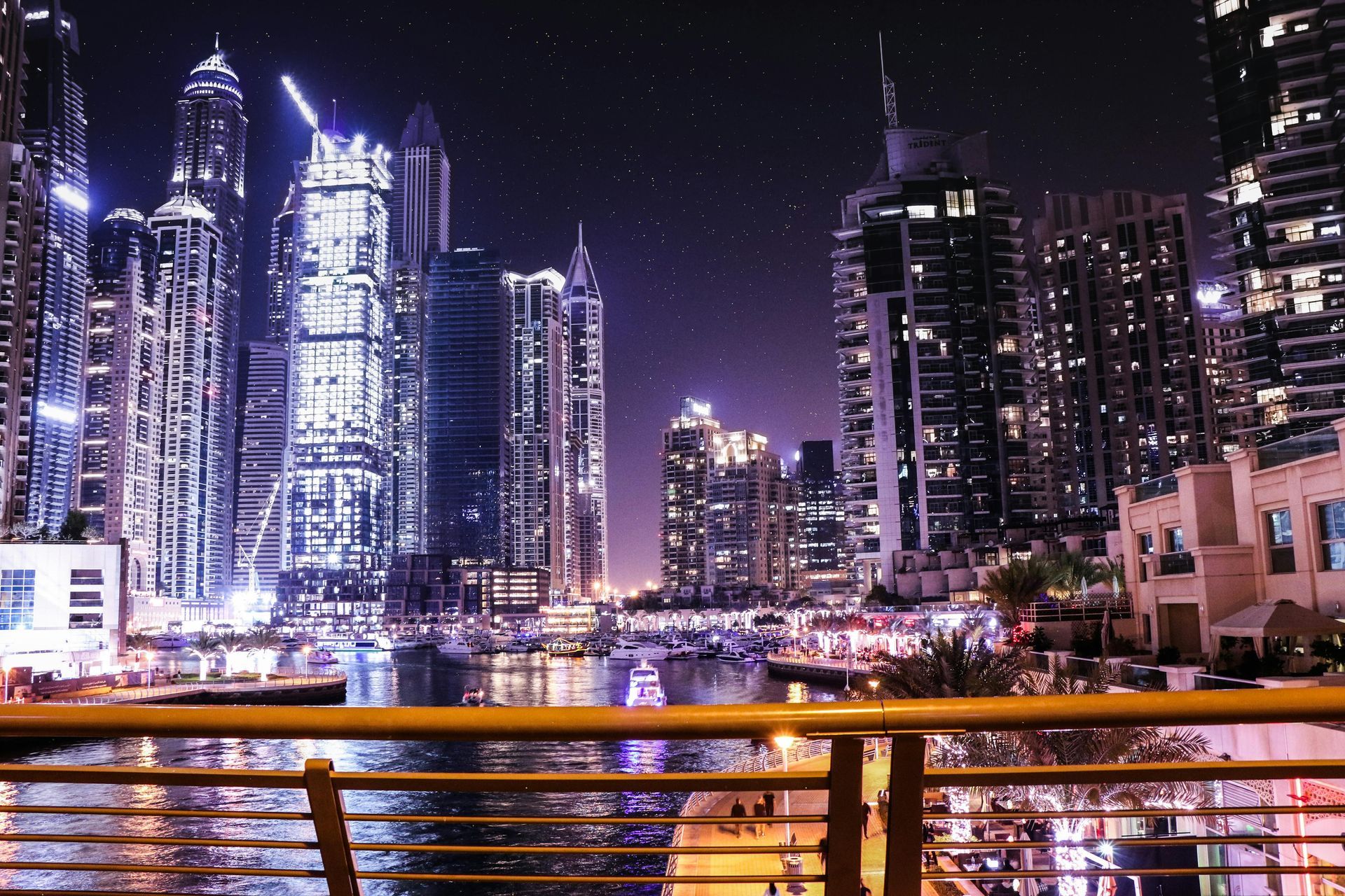 Nighttime view of a cityscape with tall illuminated skyscrapers, reflecting in the water, with a low yellow railing in the foreground.