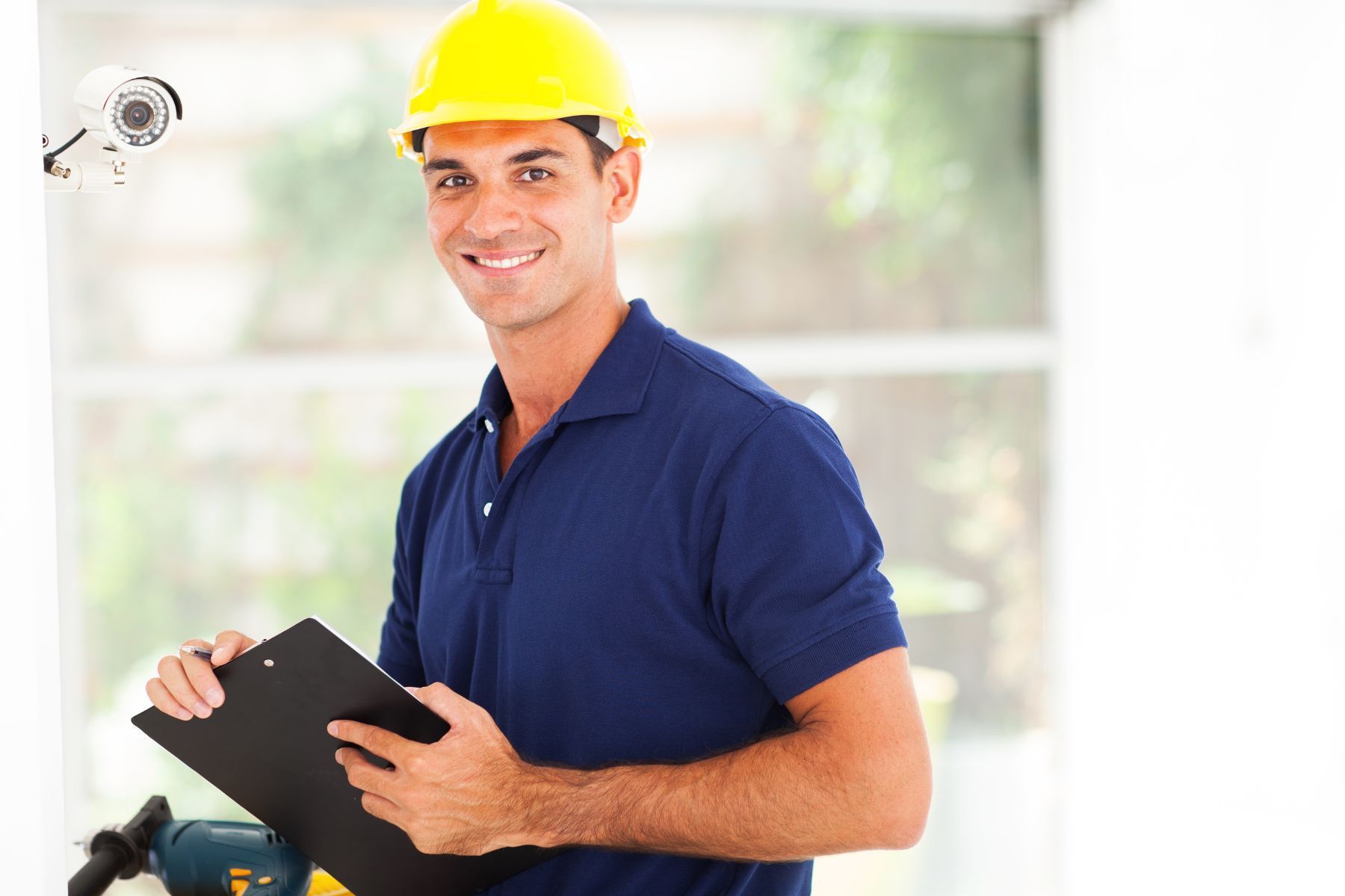 Un uomo con un casco giallo sorride, tenendo in mano un blocco per appunti vicino a una telecamera di sicurezza e a un trapano.