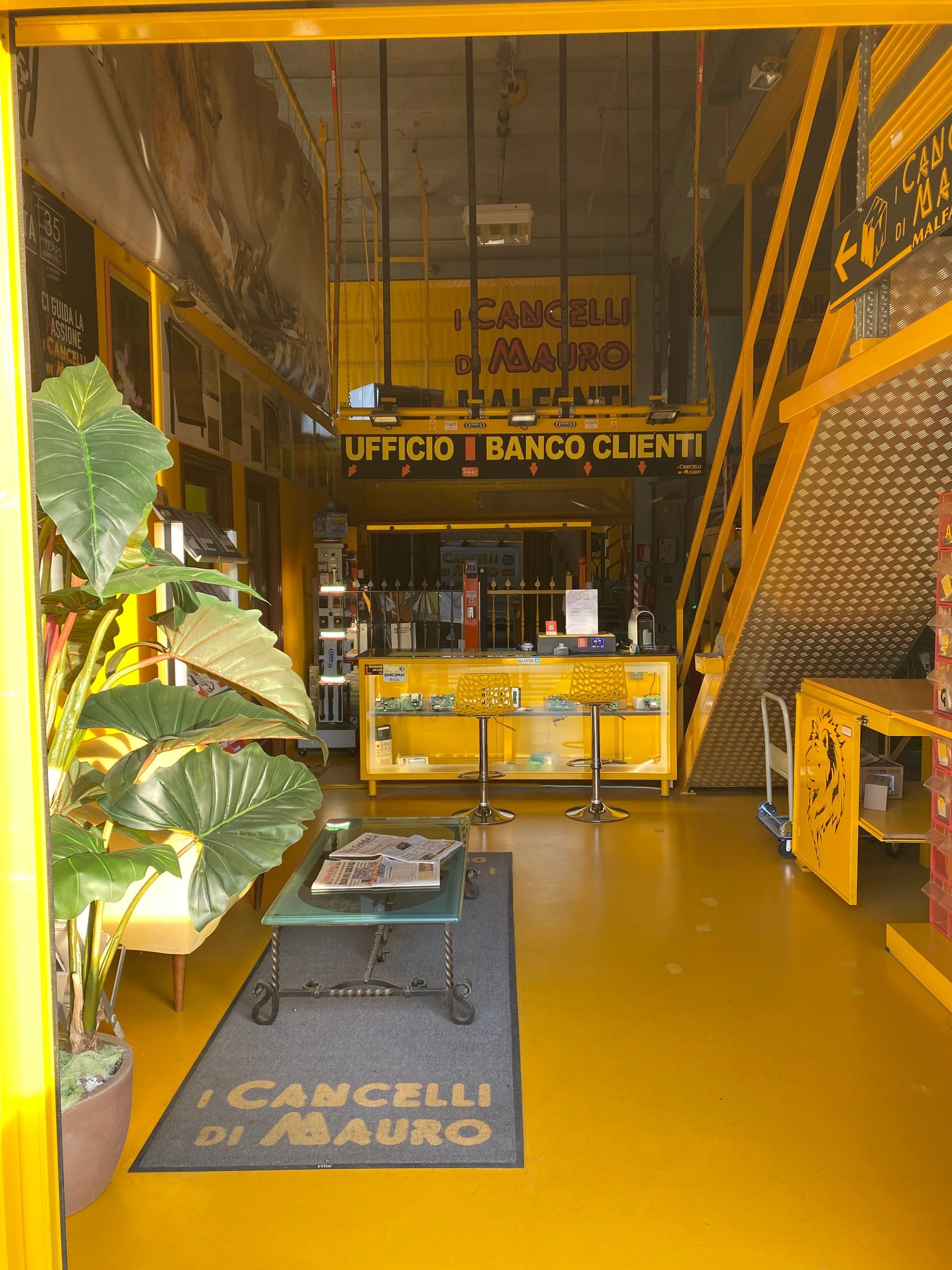 Interno giallo con area dedicata al servizio clienti, una scala e una grande pianta. Insegna: