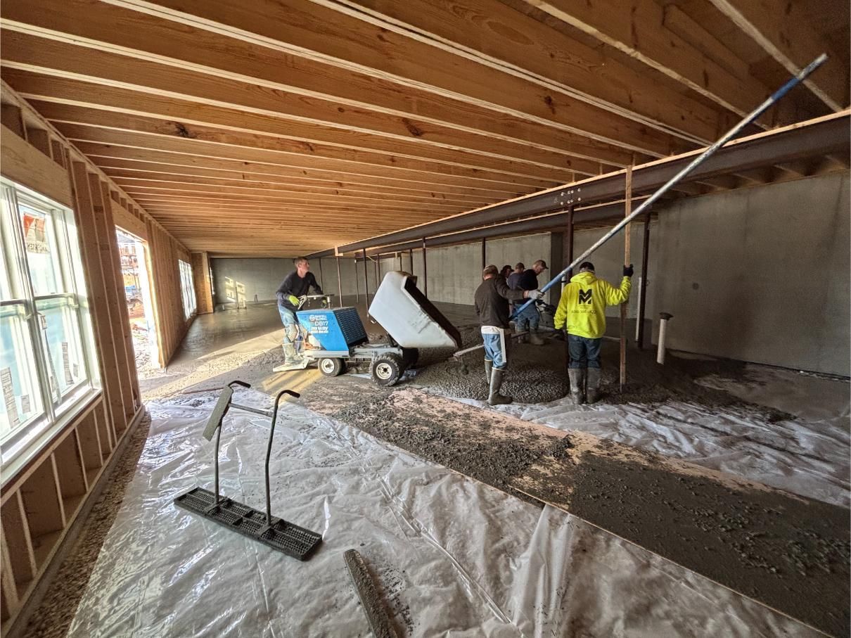 Construction workers pouring concrete inside a building with wooden beams, using a dumper and leveling tools.