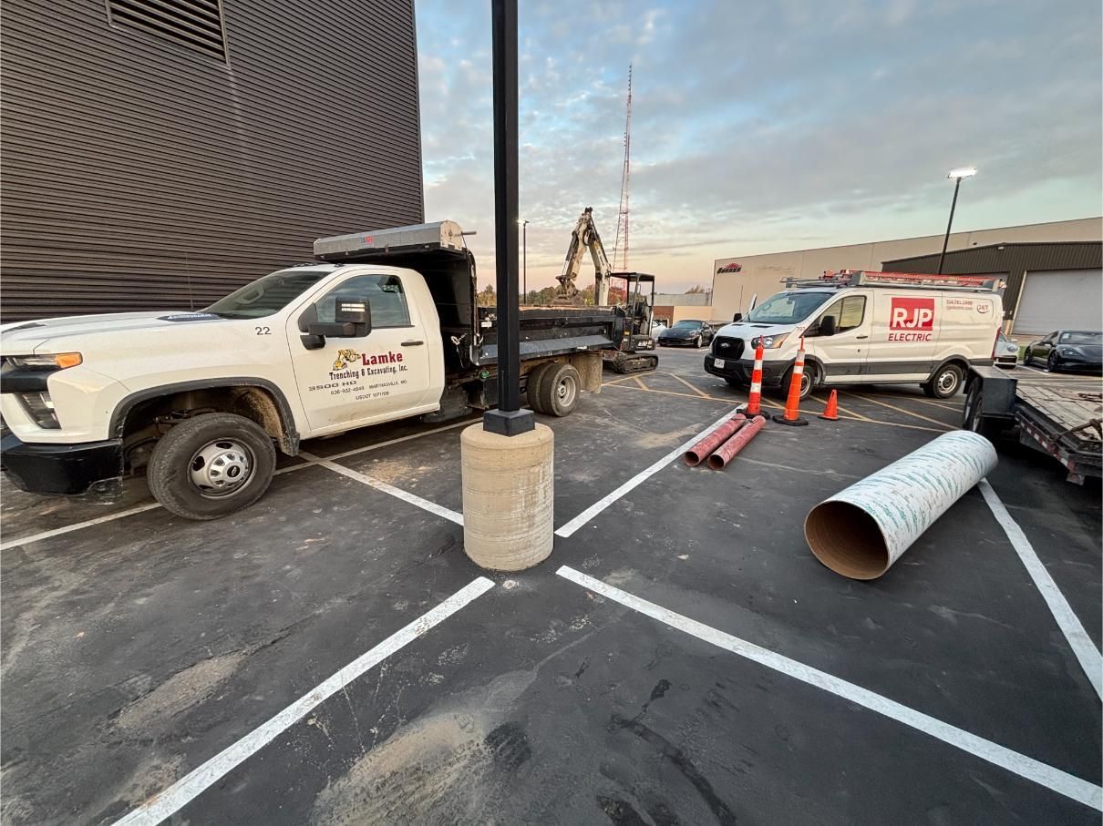 White work trucks, construction equipment, and materials in a parking lot; a pole in the foreground.