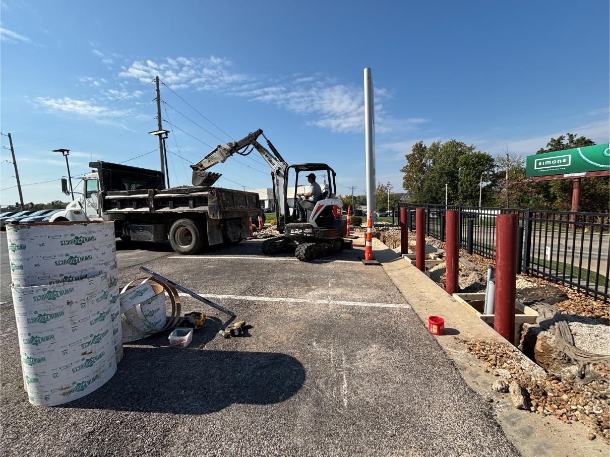 Construction site with truck, excavator, and barrier posts. Materials and equipment on asphalt under blue sky.