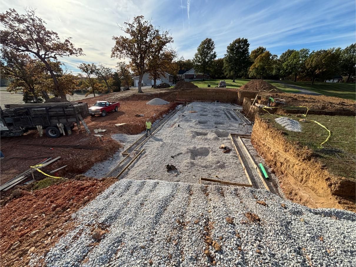 Construction site with gravel foundation and surrounding dirt. A red truck sits nearby. Sunny day.