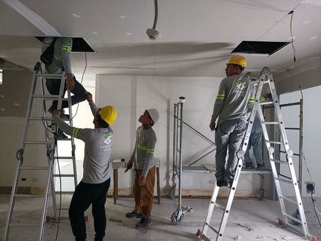 Construction workers installing electrical wiring in a white-walled room, working from ladders.
