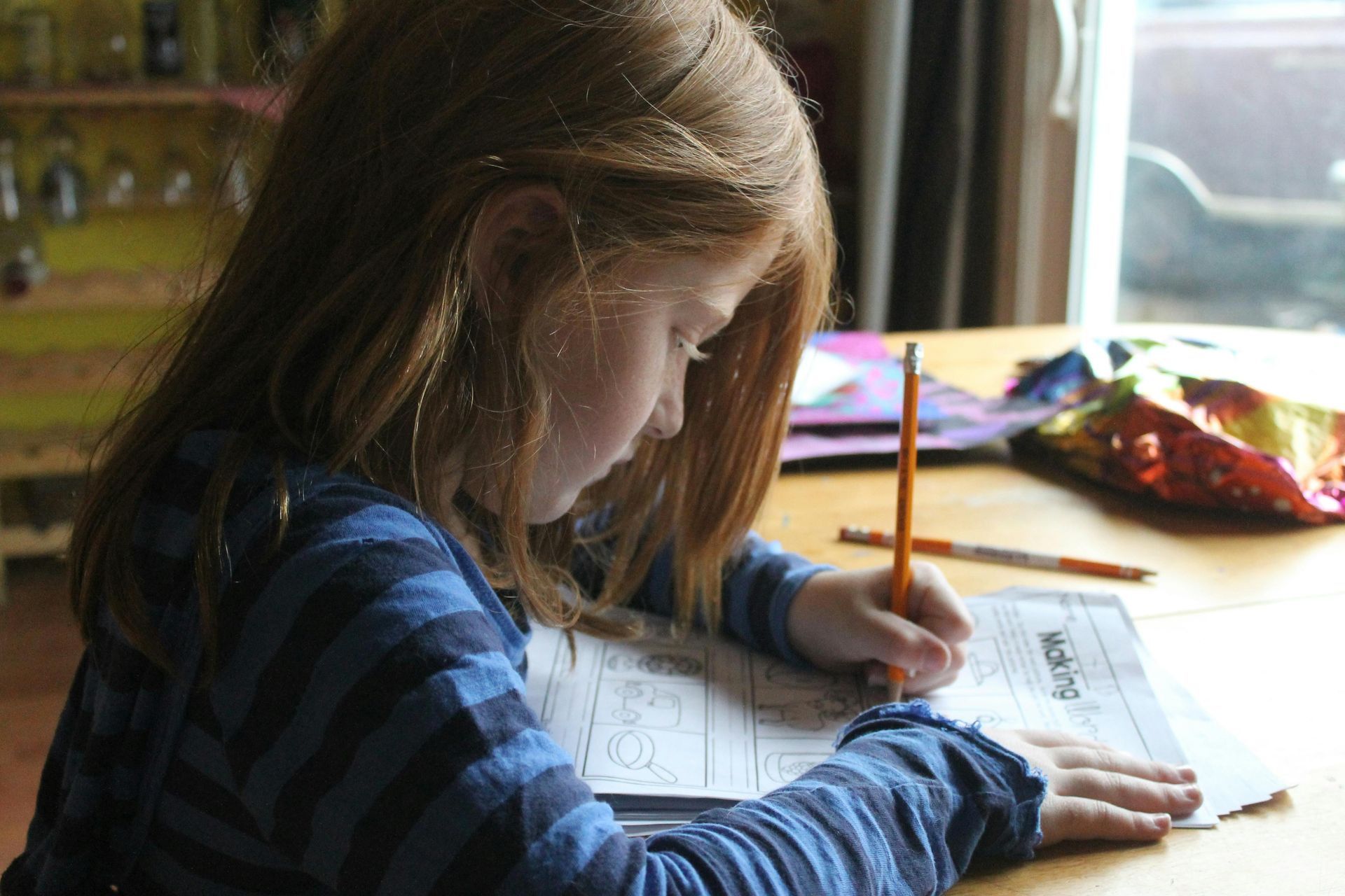 Girl with long auburn hair writing with a pencil at a table, focused on a worksheet.
