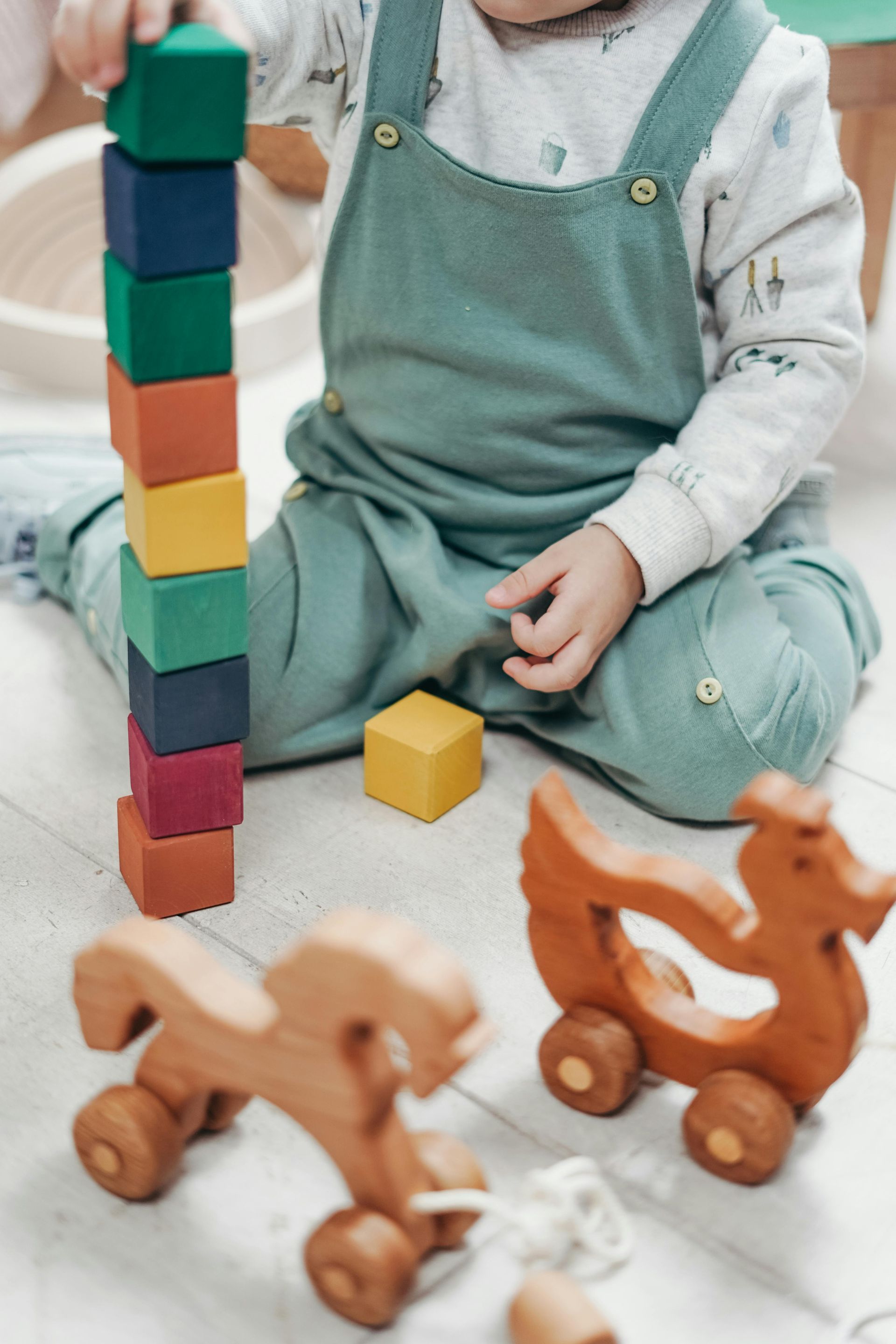 Child building a tower of colorful blocks while seated on the floor with wooden toys.