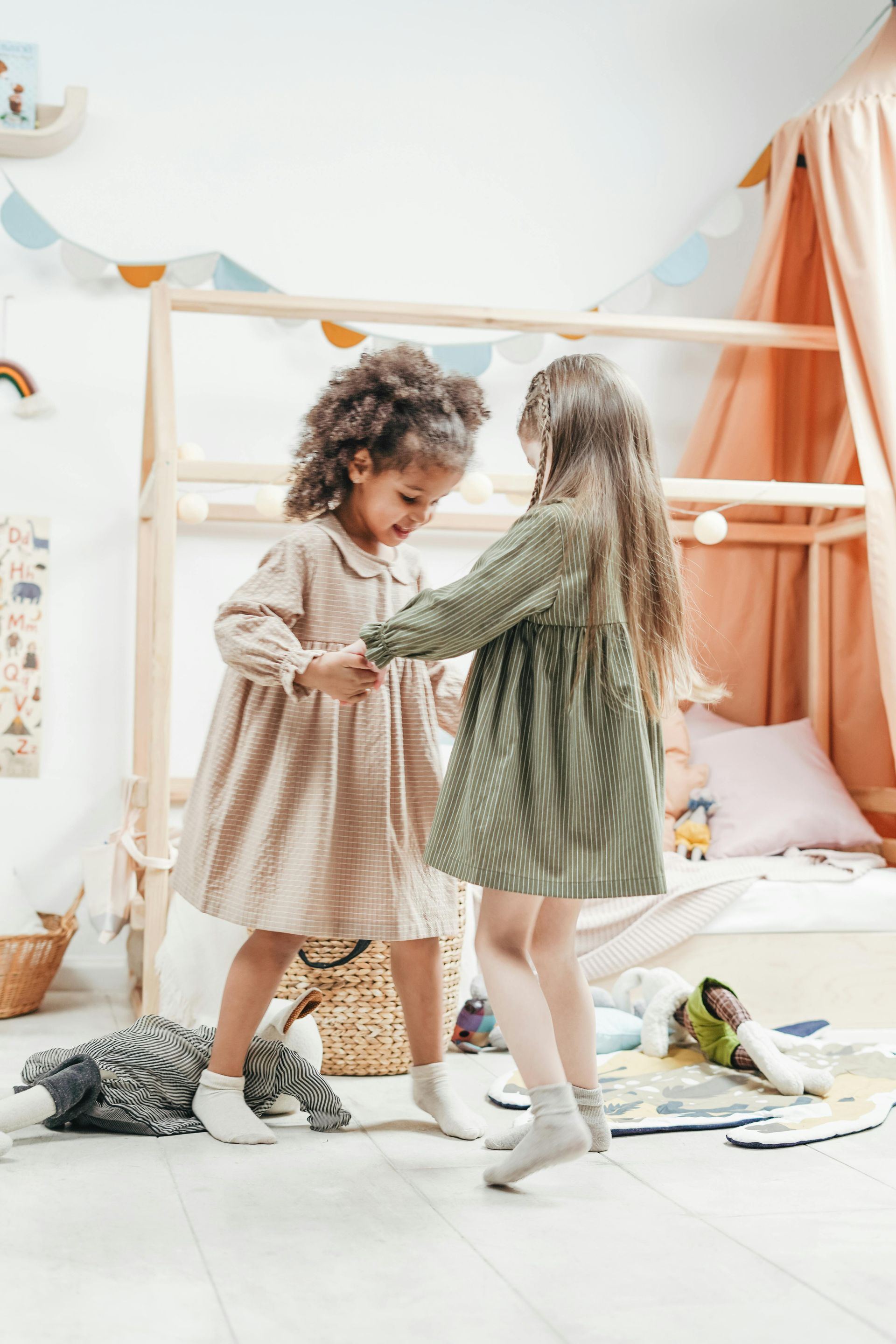 Two girls in dresses hold hands, dancing in a light-colored playroom with a canopy bed.