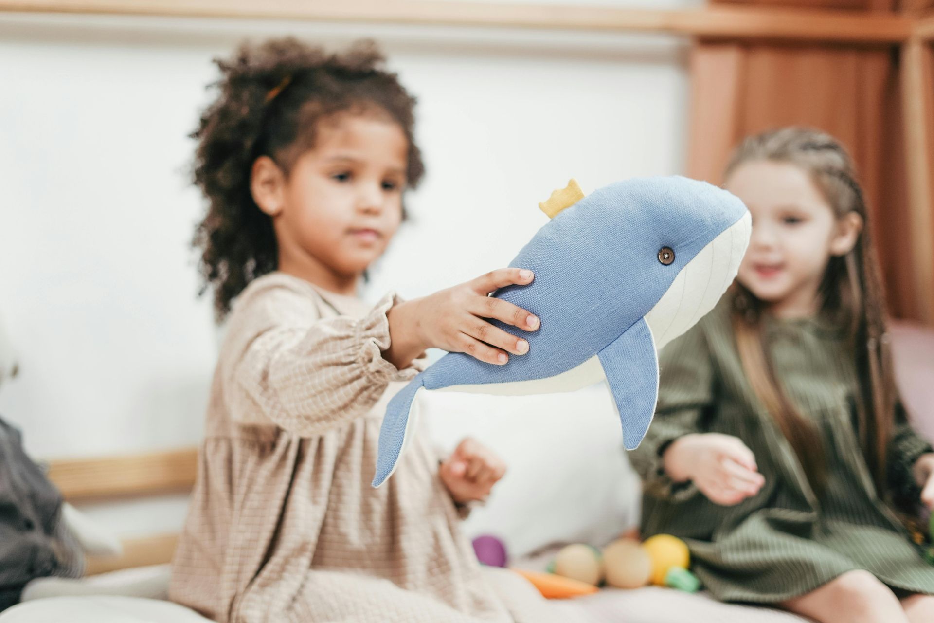 Young child holds a blue whale stuffed animal; another child smiles in the background.