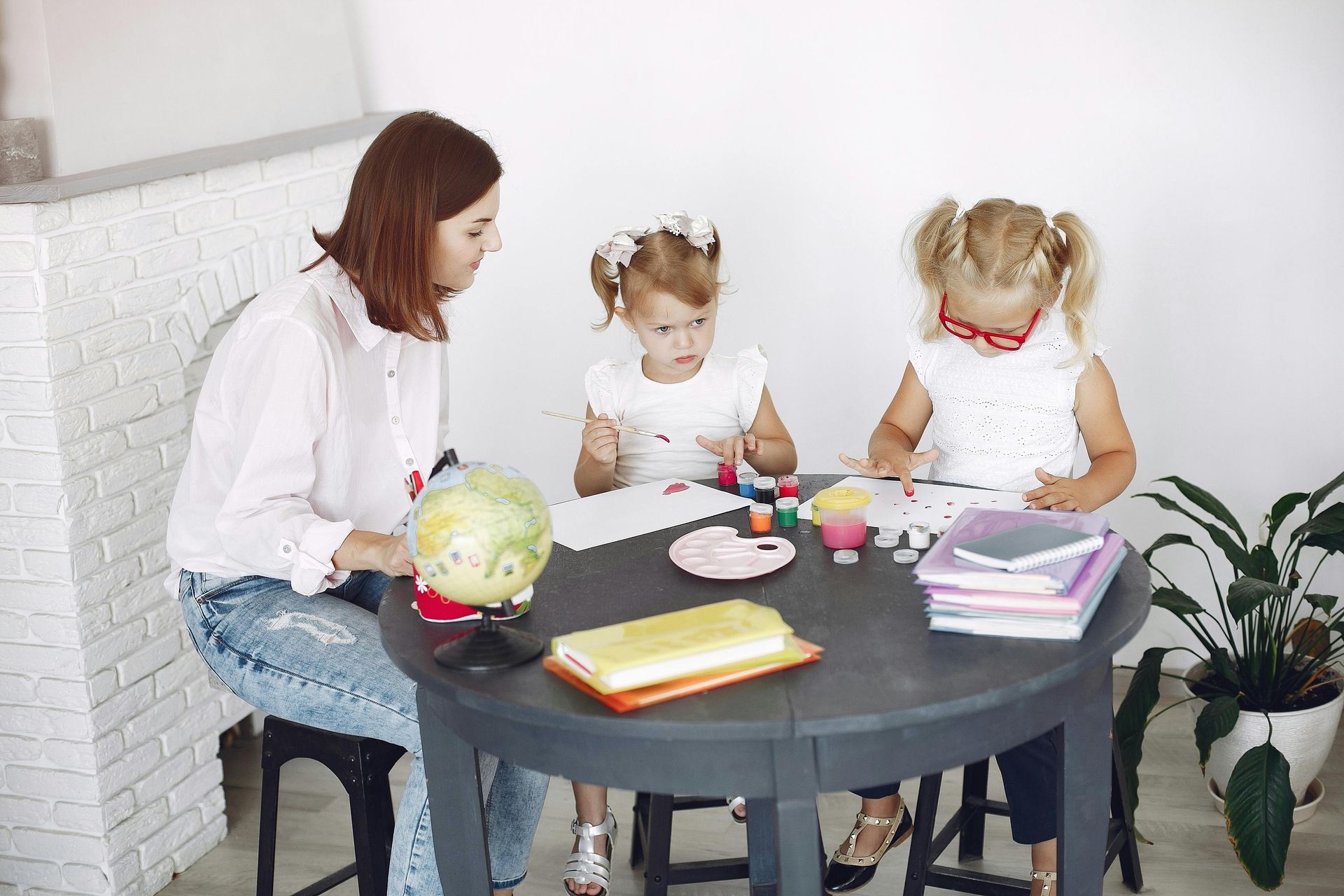 Woman helping two children seated at a round table, painting with various supplies.