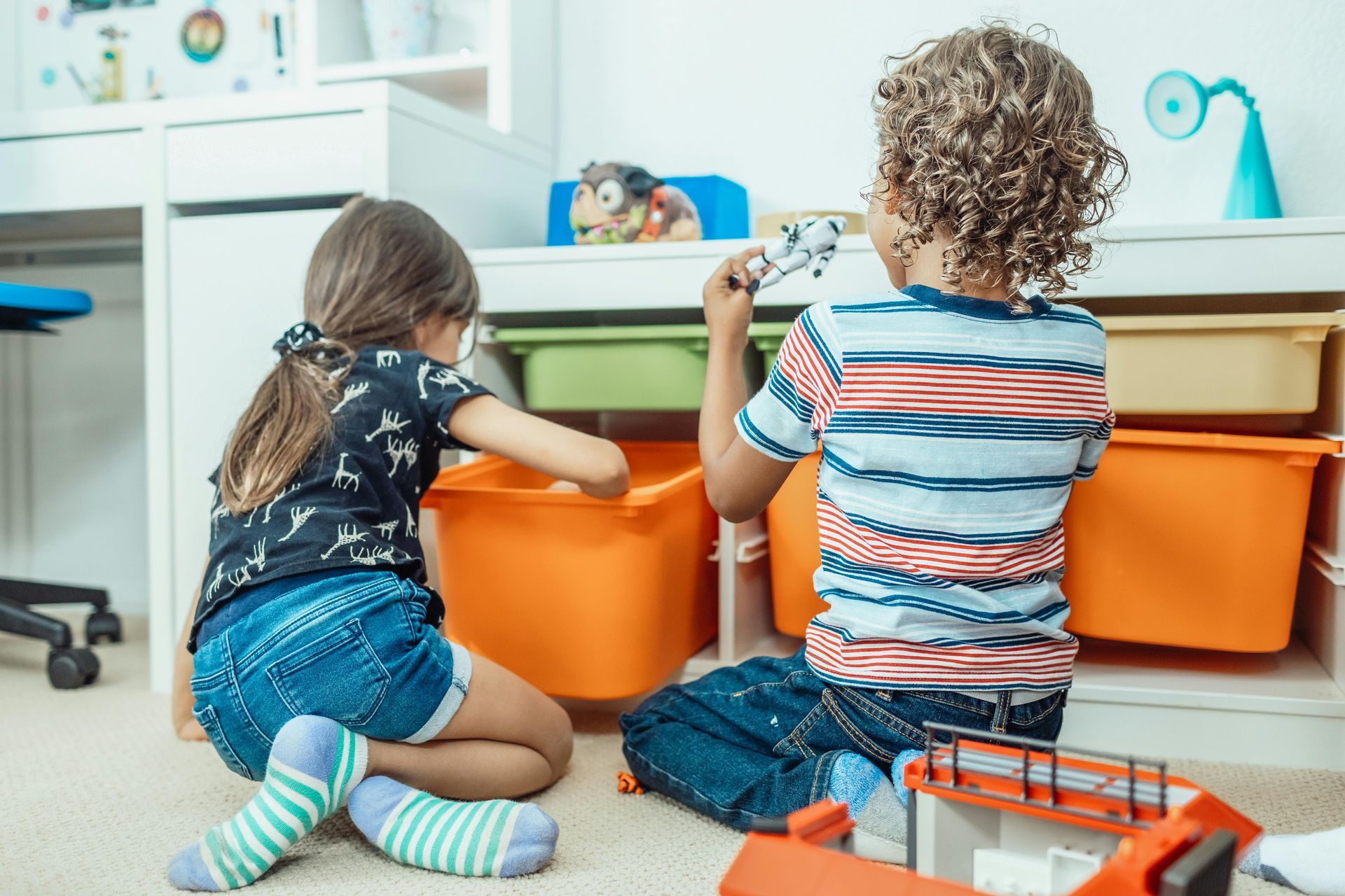 Two children playing with toys in front of orange bins in a room.