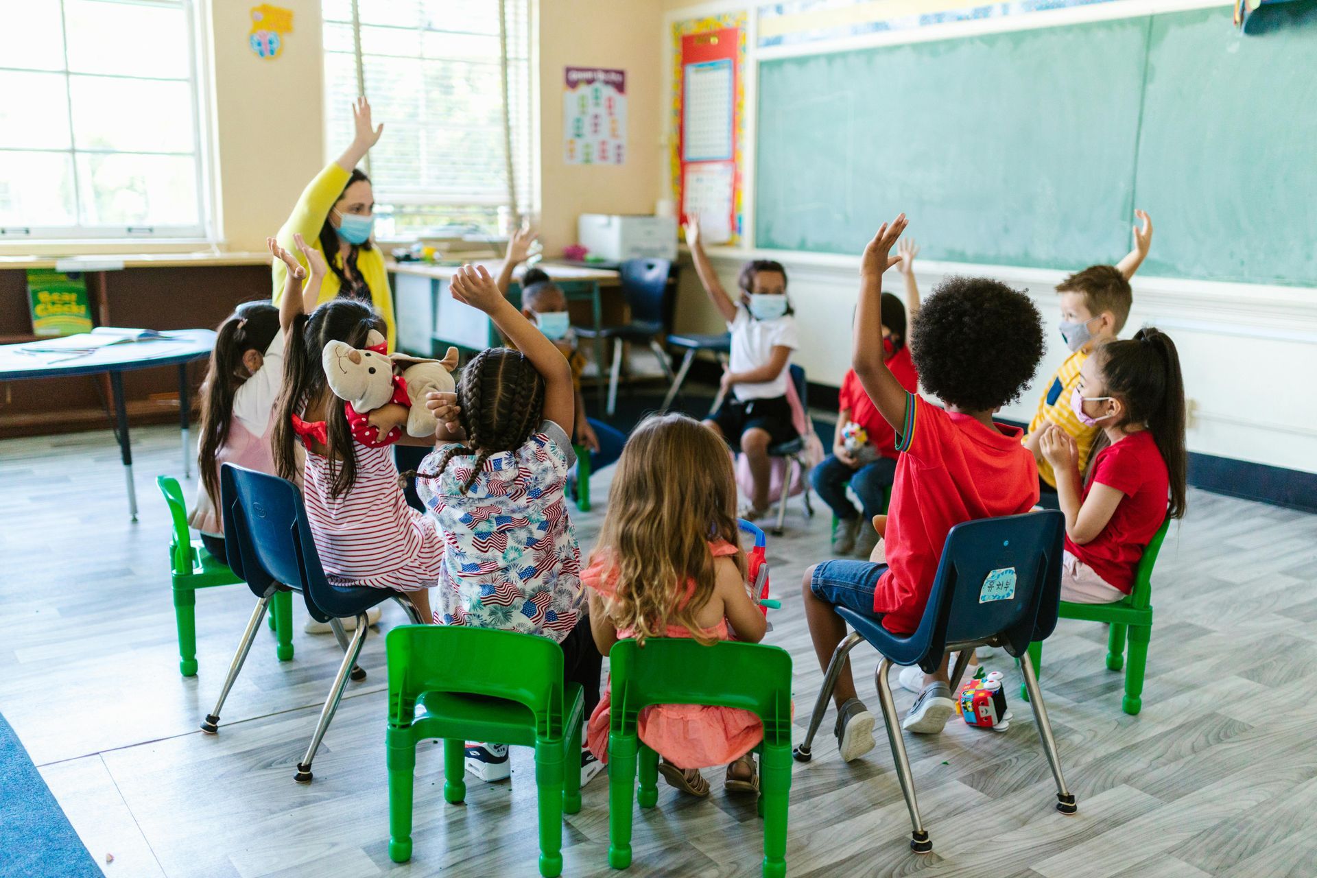 Children and teacher in masks sitting in a circle, raising their hands in a classroom.