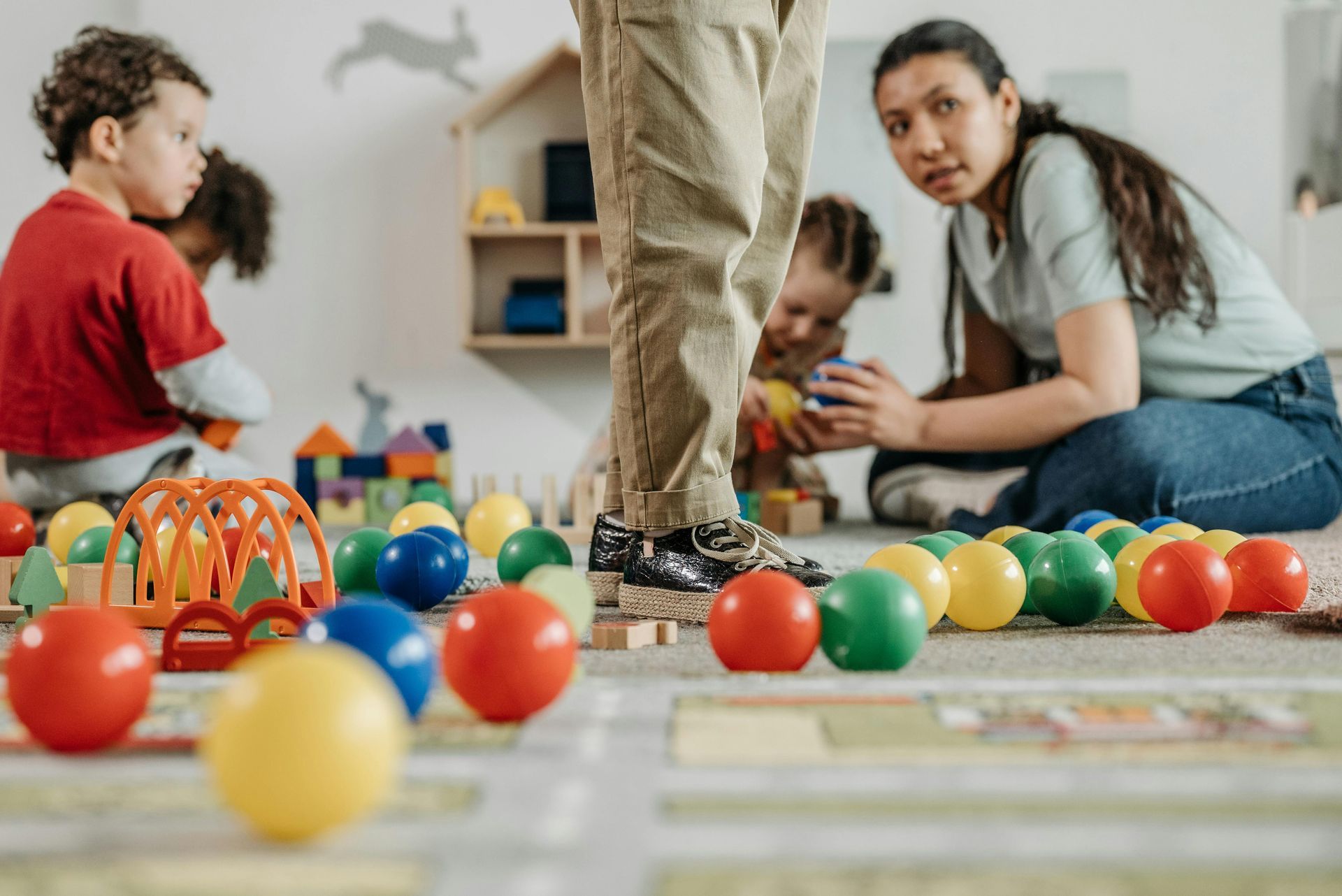 Children and caregiver playing with toys in a brightly lit playroom. Colorful balls and blocks are scattered on the floor.