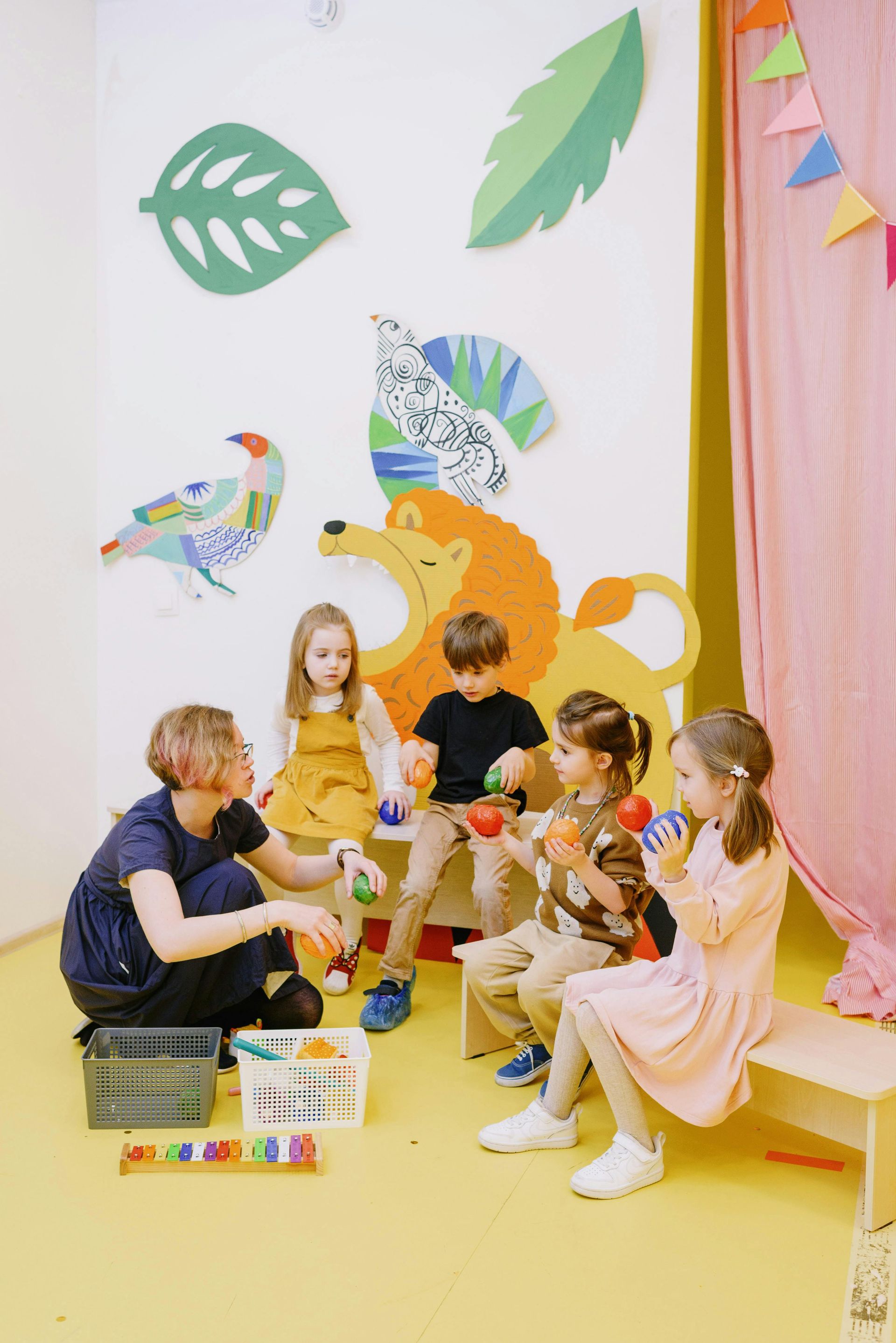 Children and teacher in a playroom with lion mural, playing with colorful balls.