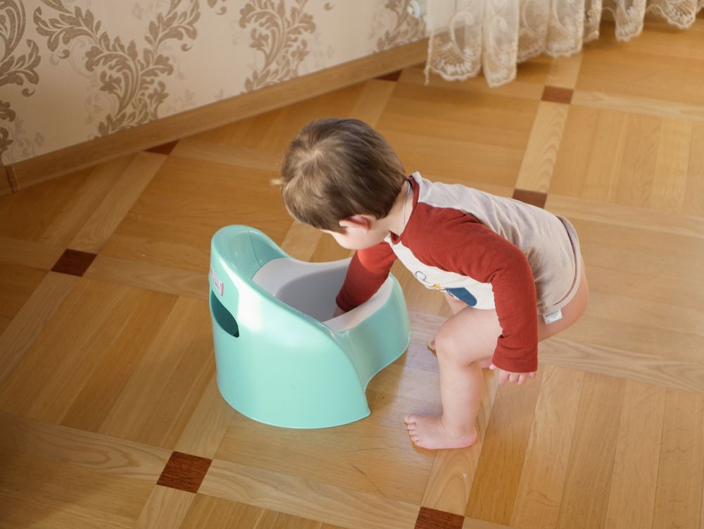 Child by a light blue potty chair on a wood floor, reaching inside.