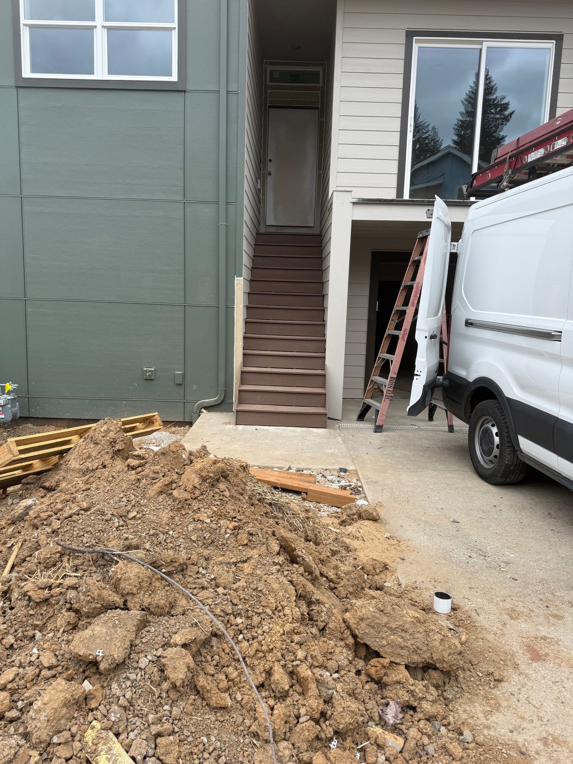 Dirt pile, wooden stairs leading to a door, van parked, ladder. Grey and green buildings.