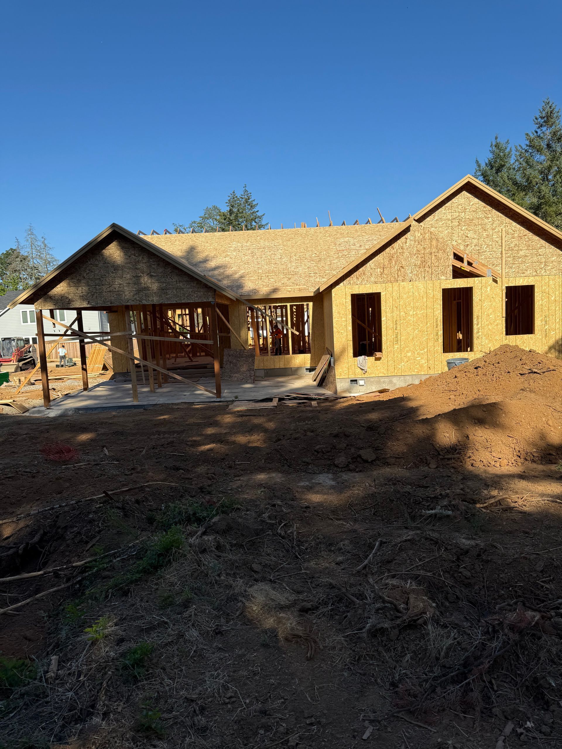 A house under construction with exposed wooden framing and a porch, on a sunny day.