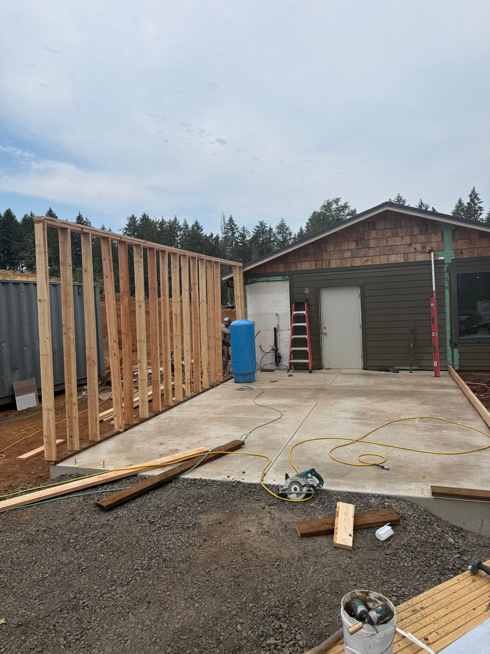 Construction site: Wood frame wall being built next to a concrete pad, a garage and container are in the background.