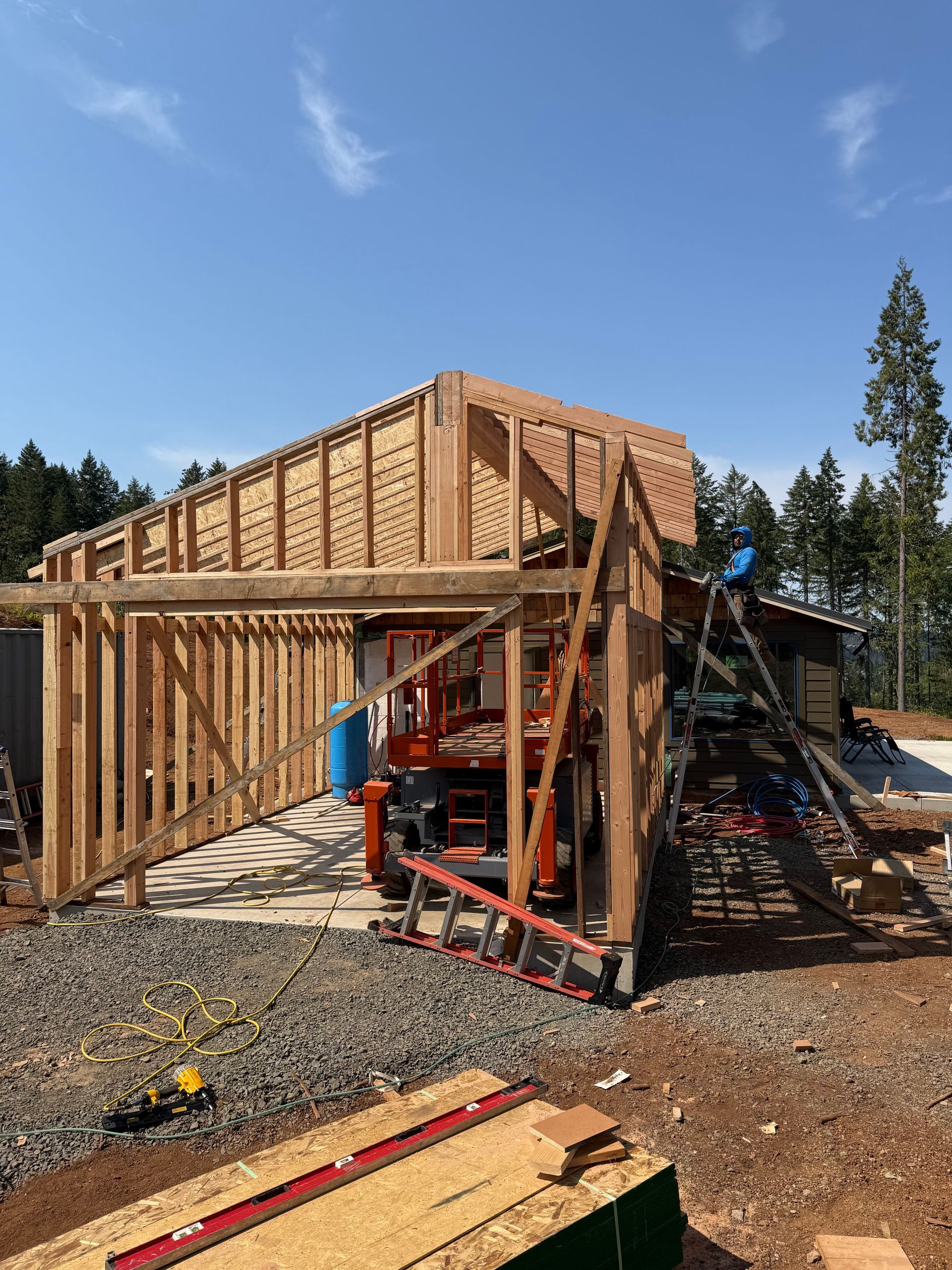 Construction of a wood-framed building with a red lift and ladder on a gravel lot under a blue sky.