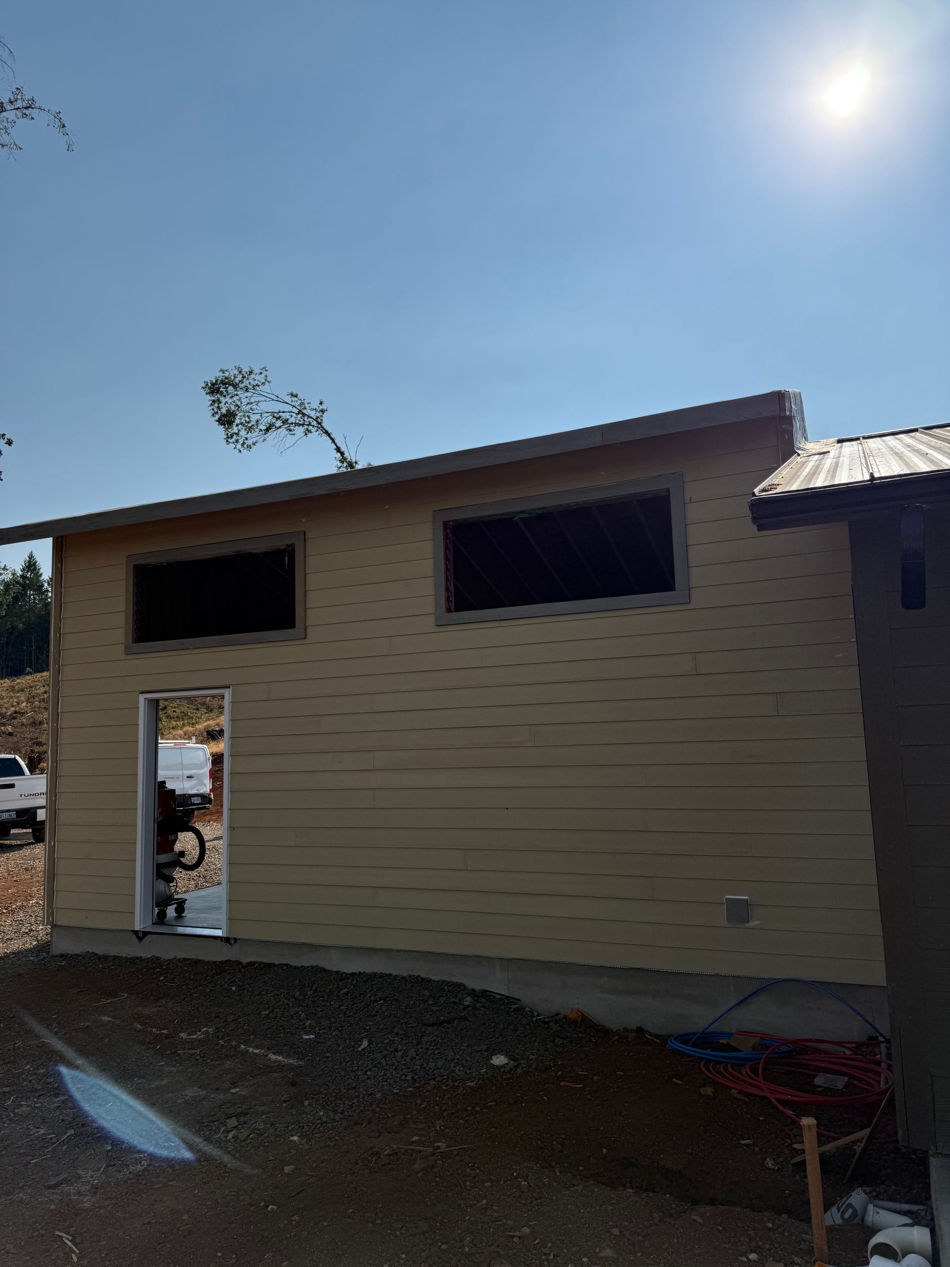 Exterior view of a building under construction. Tan siding, open door and window frames, bright sun.