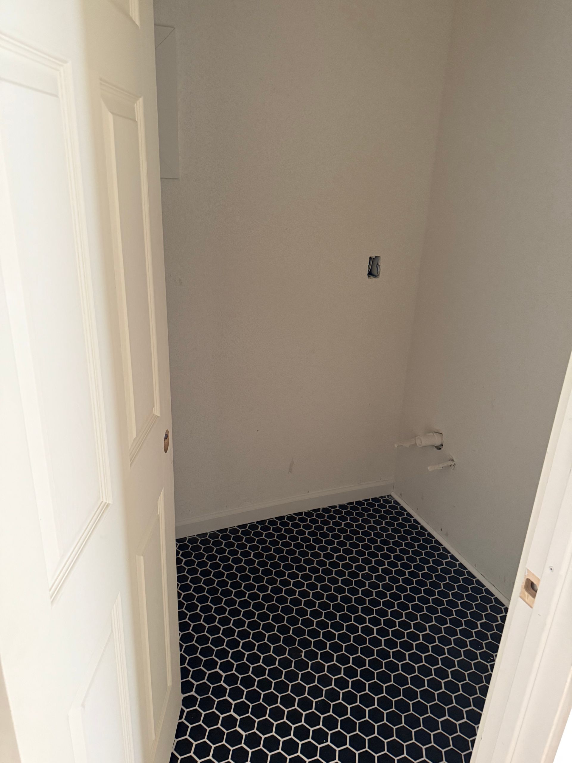 Small laundry room with black hexagon tile floor and white walls. Open white door in the foreground.