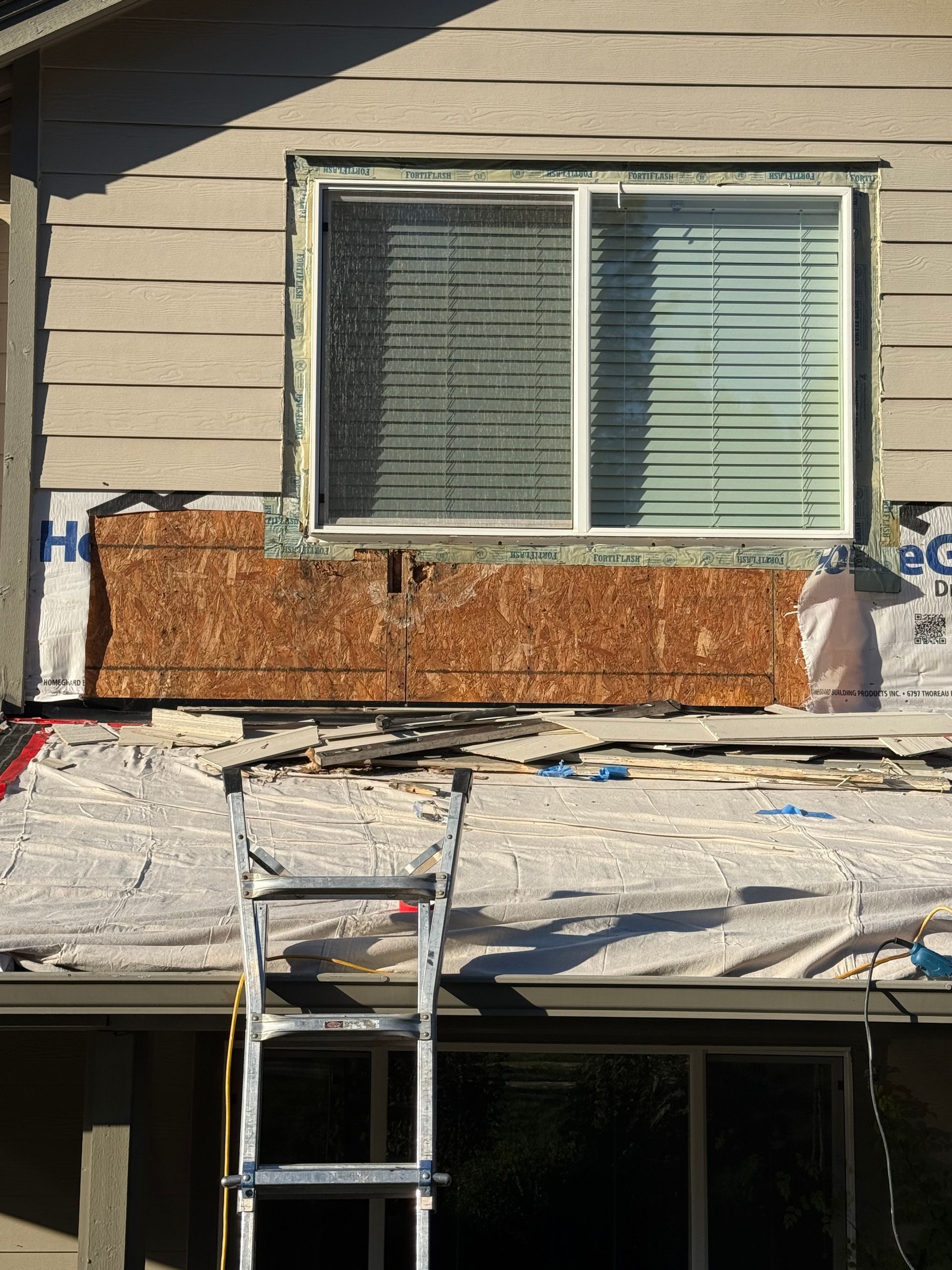 Window frame being worked on, exposing wood framing and sheathing, ladder below.