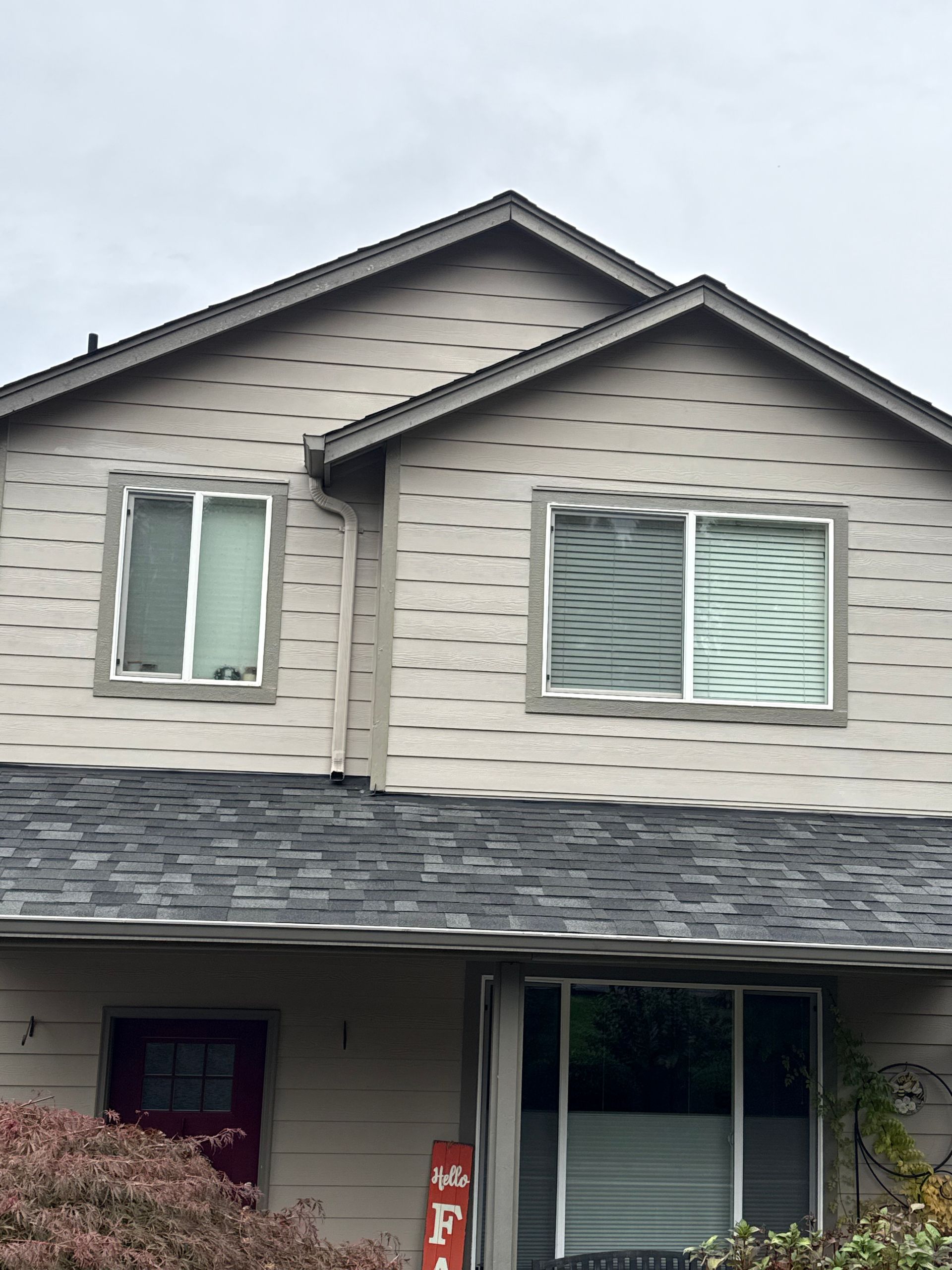 Two-story house with tan siding, gray roof, white-framed windows, and red door. Overcast sky.