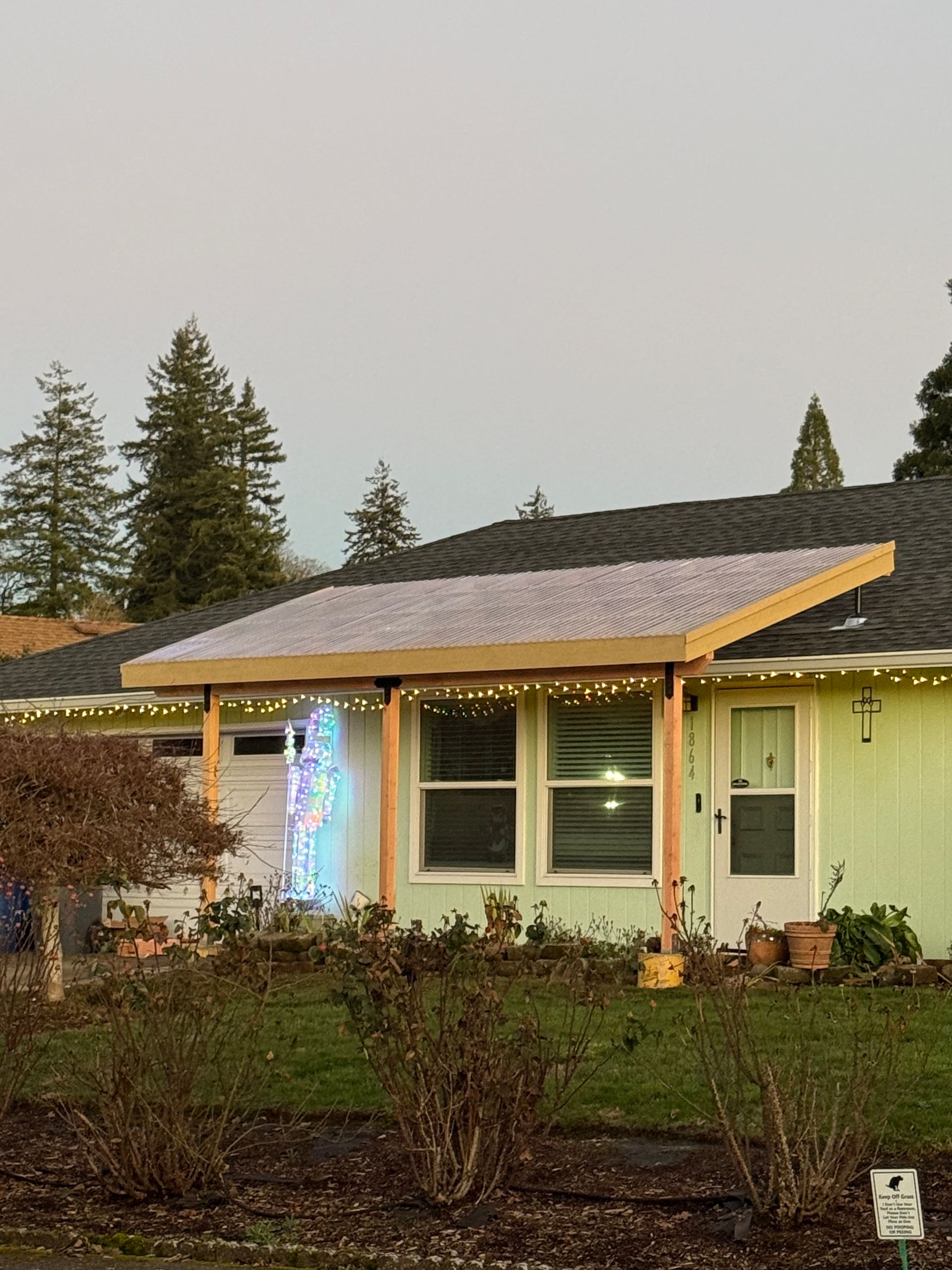 A light green house with a covered porch decorated with string lights, trees in background.