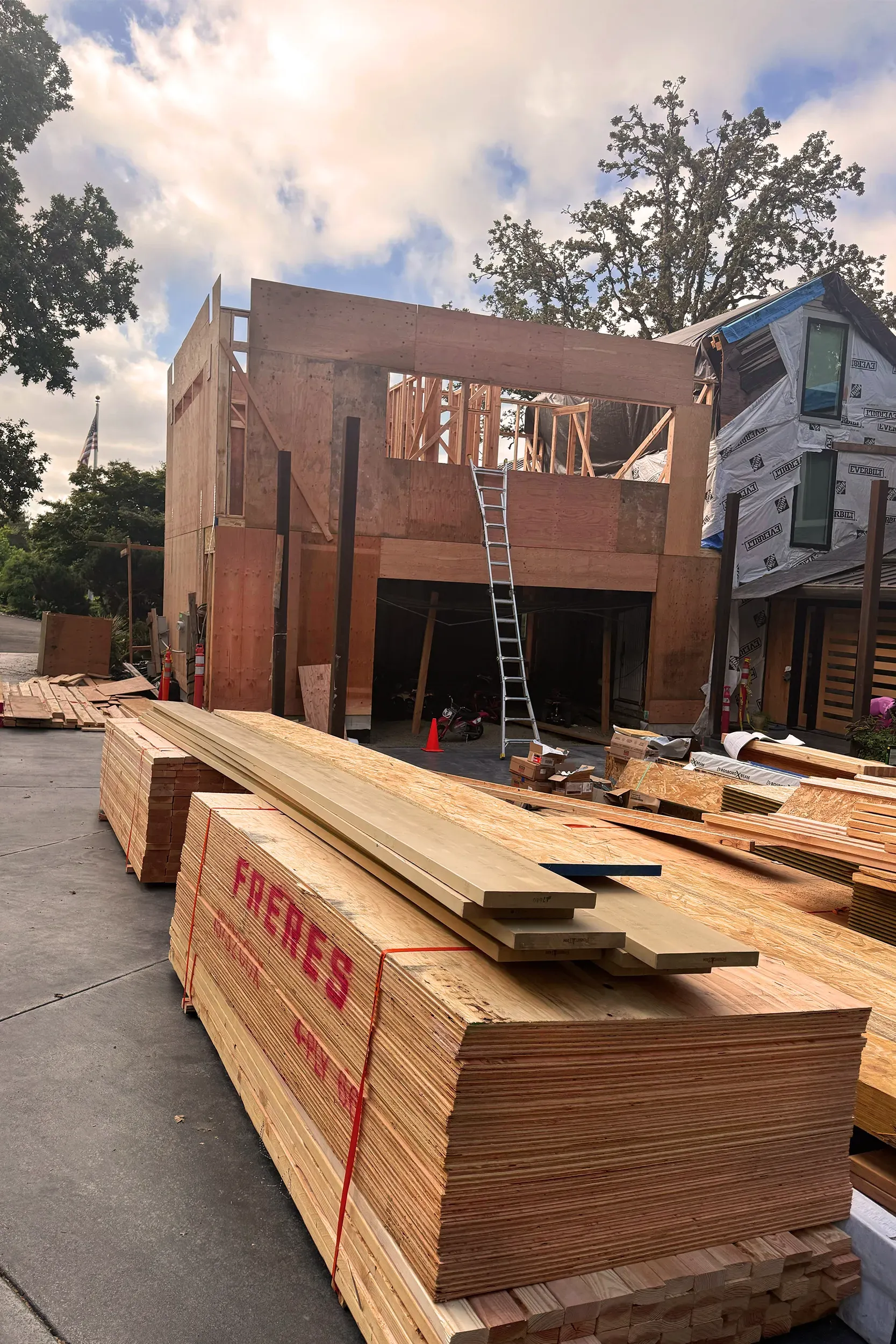 Construction site: wooden building frame with stacked lumber in foreground, ladder, and cloudy sky.