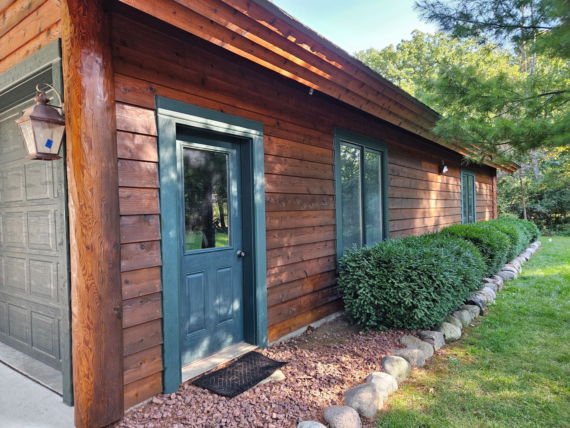 Brown log cabin with green trim, door, windows, and garage door. Green bushes line the front.